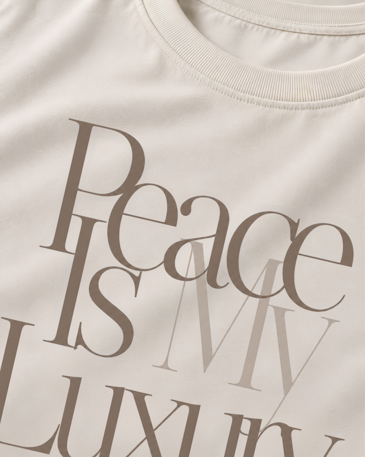 Close-up of a cream-colored crewneck t-shirt featuring a layered graphic print of the words 'Peace IS MY Luxury' in muted taupe-brown typography.