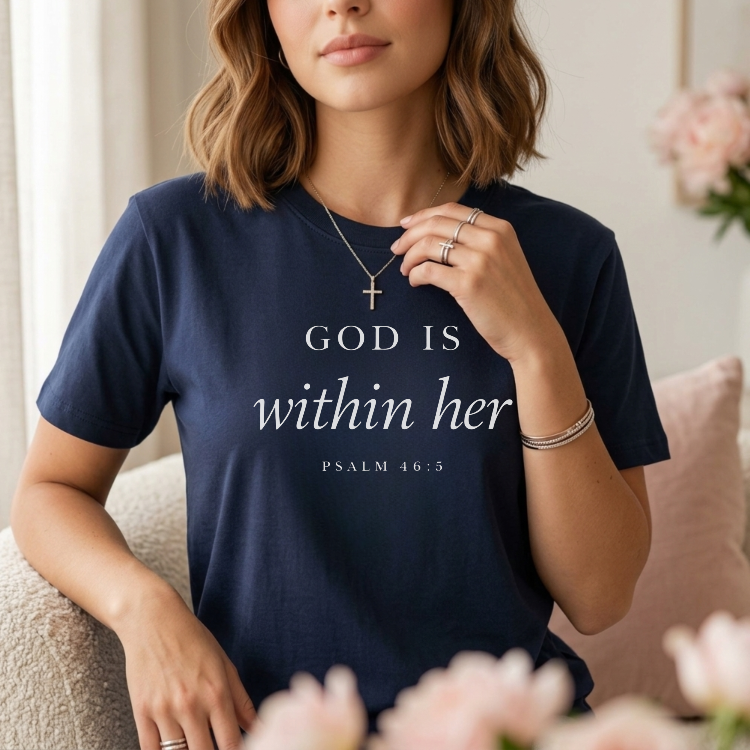 God is within her Psalm 46:5 bible verse on navy T-shirt. Wearing gold cross necklace. Christian woman standing in front of flowers and a window wearing a Scripture Tee.