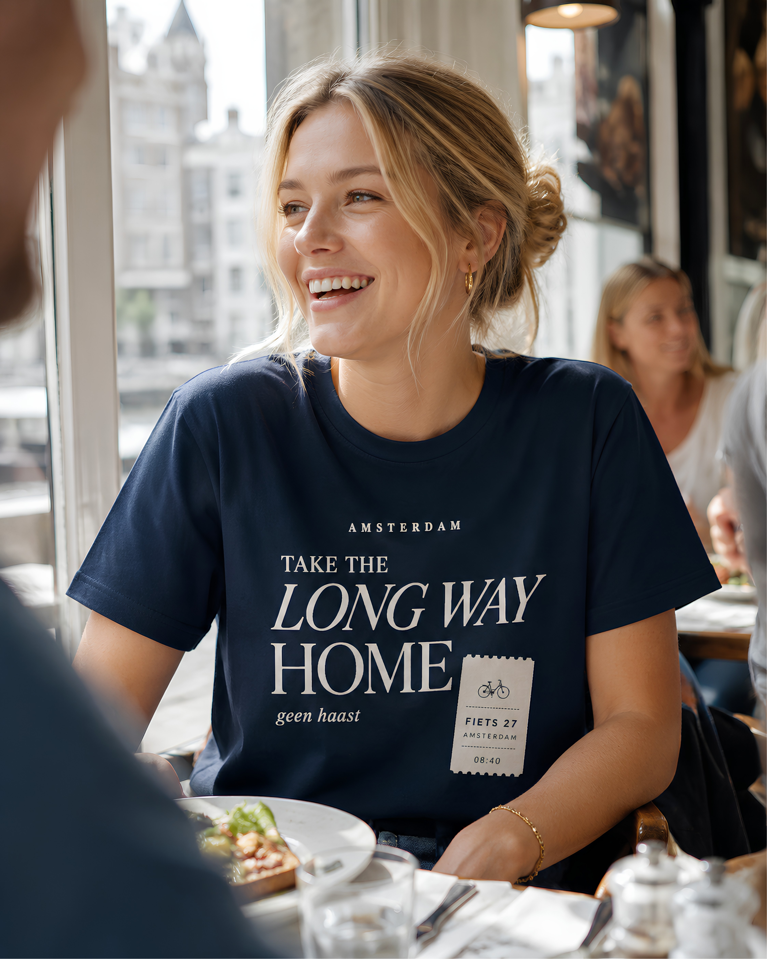 Woman wearing navy Amsterdam Take the Long Way Home T-shirt while enjoying lunch at a café in Amsterdam