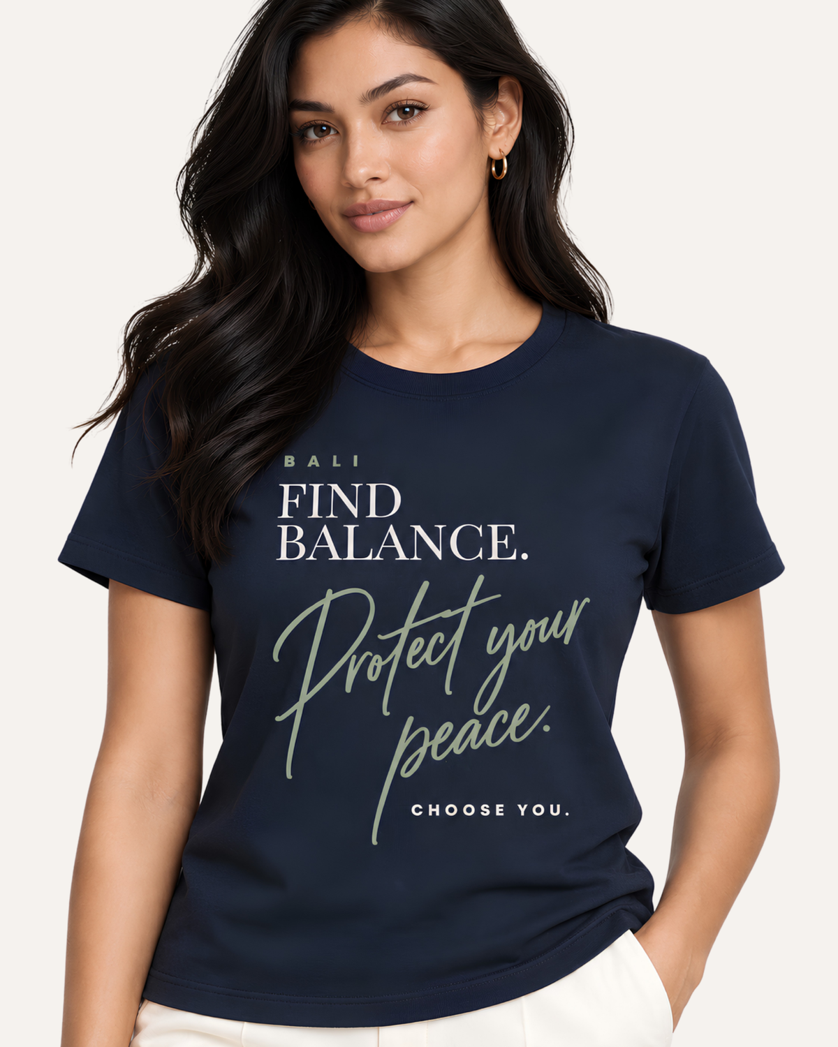 A woman wearing a navy blue graphic t-shirt with white and sage green text that reads 'BALI FIND BALANCE. Protect your peace.