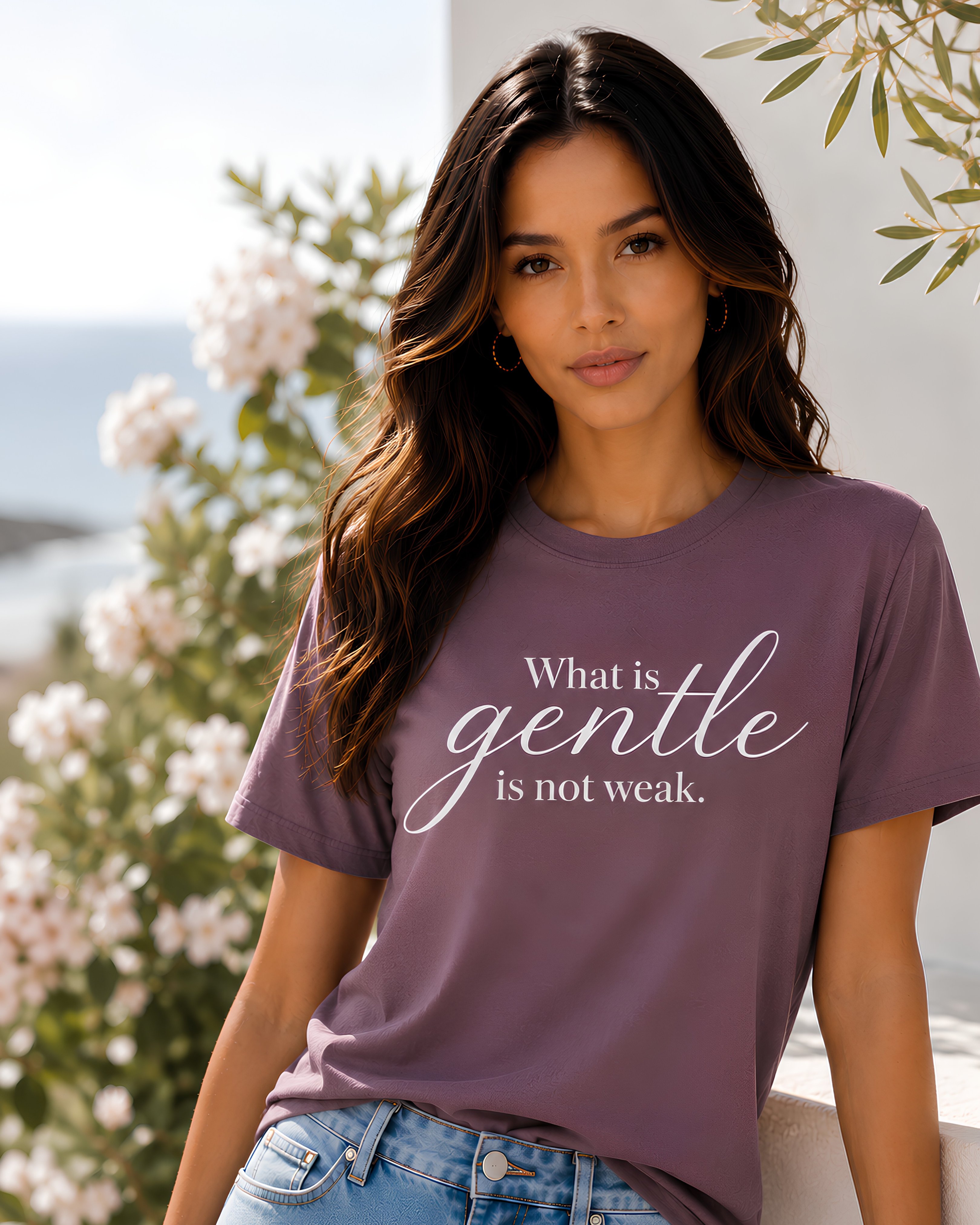 Woman wearing the What Is Gentle Is Not Weak Christian T-shirt for women, inspired by Galatians 5:22–23 and biblical gentleness. A mauve-purple graphic t-shirt paired with blue jeans, set against a soft-focus background of white flowers and green leaves.