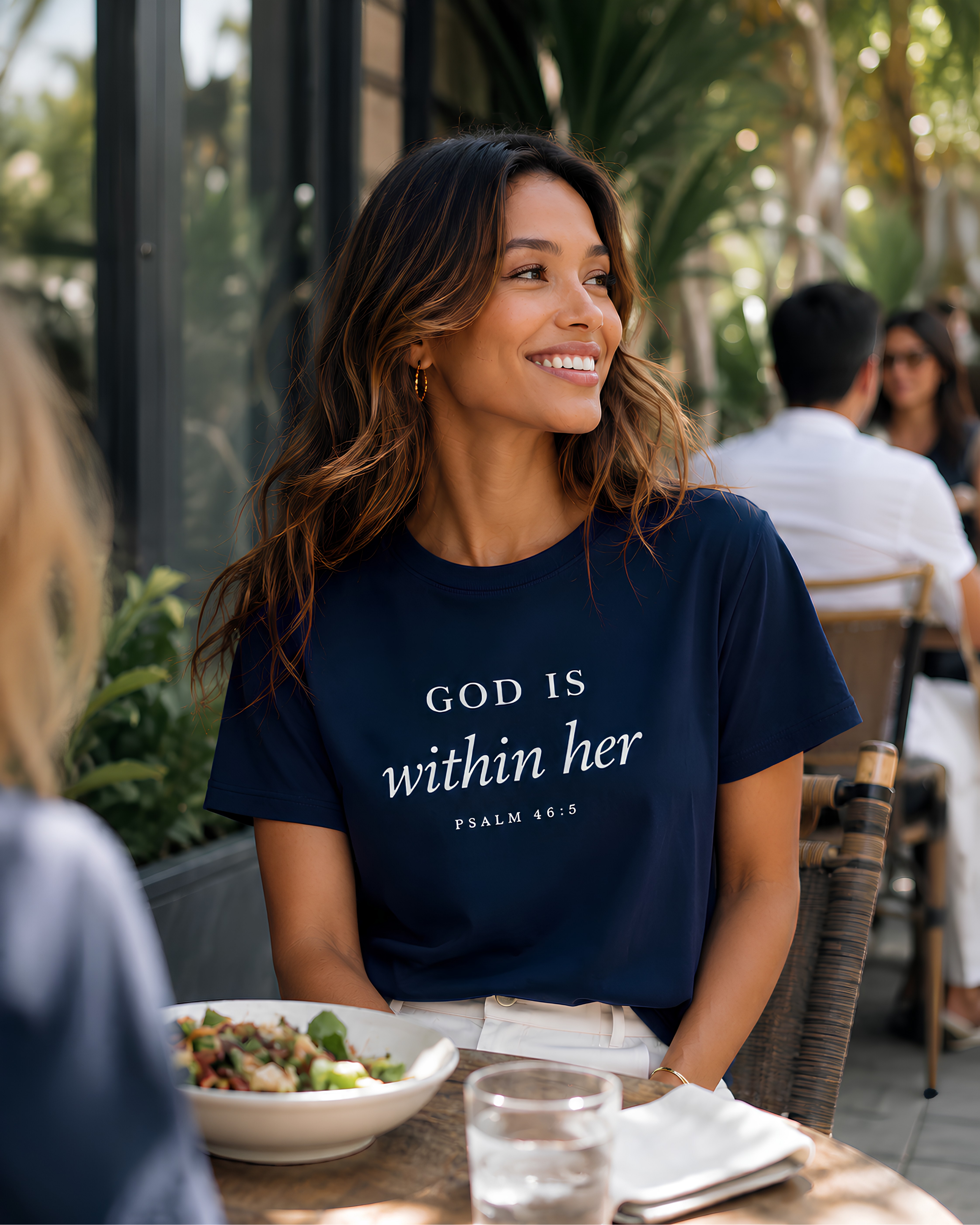 Woman wearing a navy God Is Within Her Psalm 46:5 Christian tee enjoying lunch with friends at a bright outdoor café.