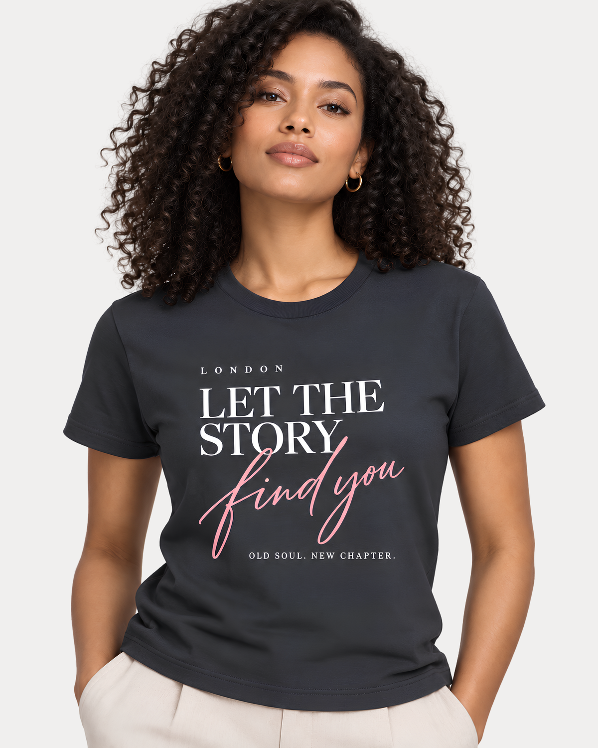 A woman with curly dark hair wearing a charcoal gray graphic t-shirt with the text 'LET THE STORY find you' in white and pink, paired with light beige trousers, against a light gray studio background.