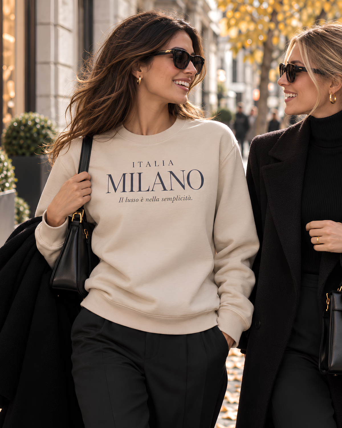 Woman wearing a beige Milano Italy sweatshirt while walking with a friend on an autumn street in Milan, styled with black tailored trousers and a coat.