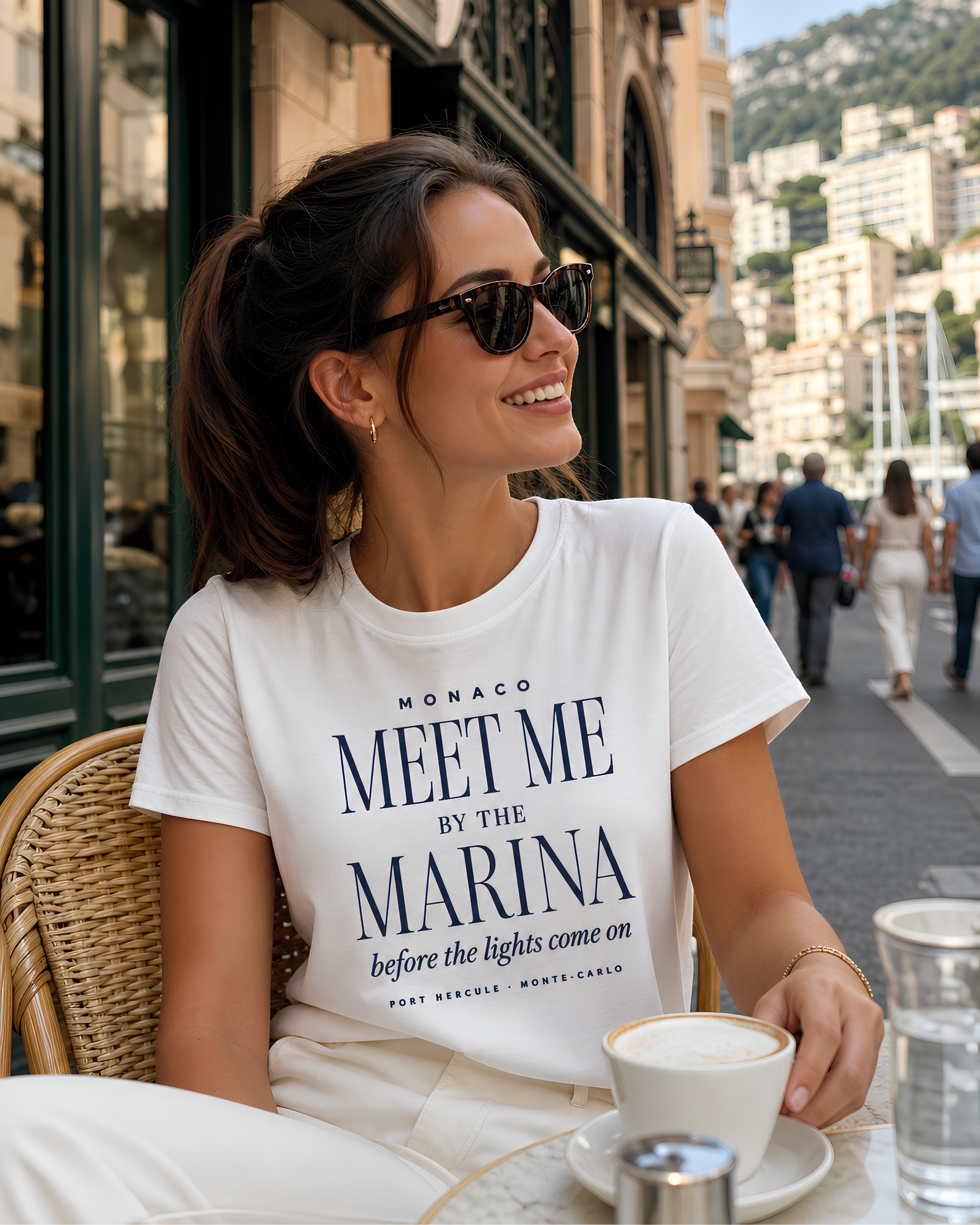 Woman wearing a white Monaco Meet Me by the Marina travel T-shirt at a café near the Monaco waterfront