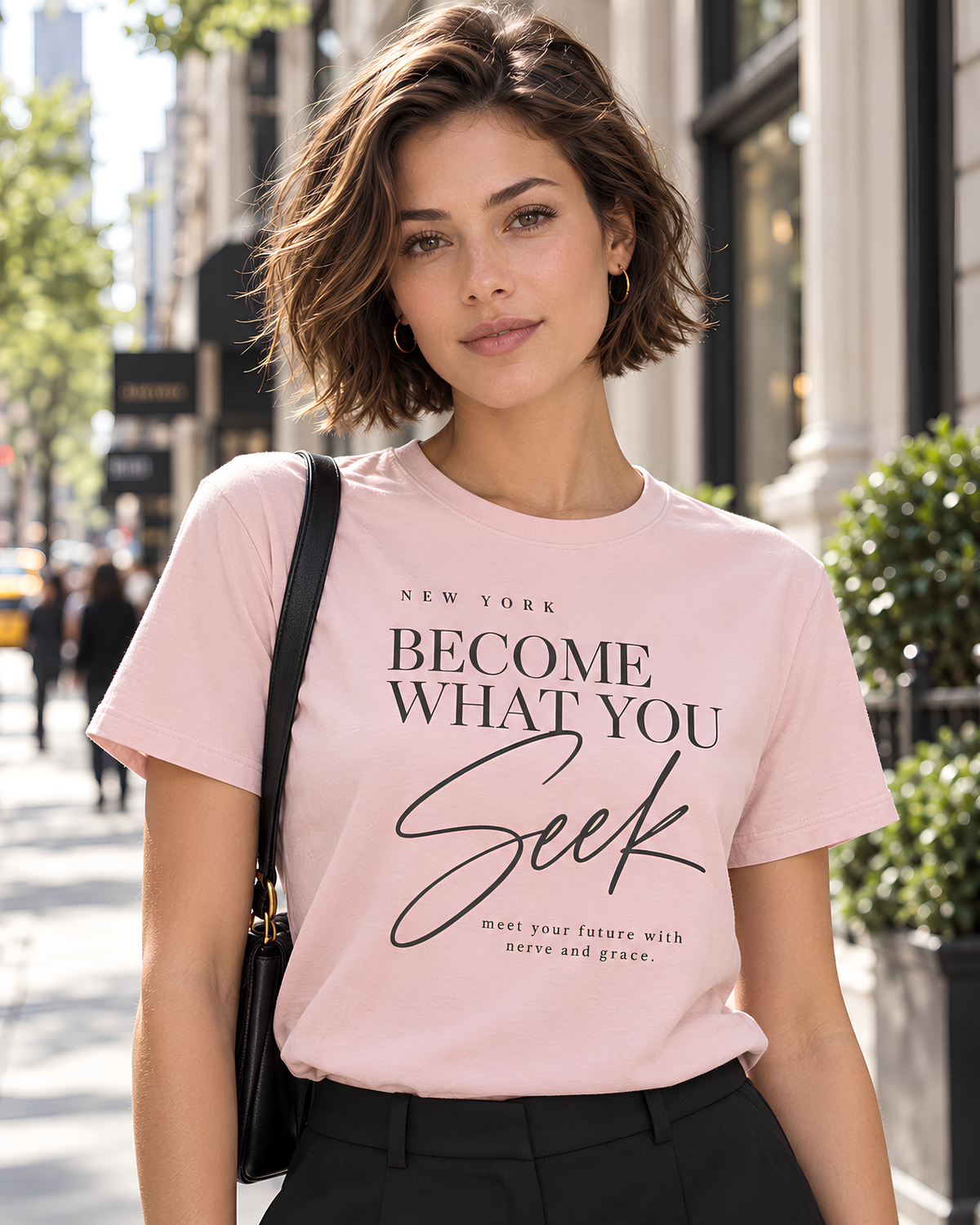 Woman wearing a pink New York graphic tee for women with “Become What You Seek” editorial typography on a bright New York City street.