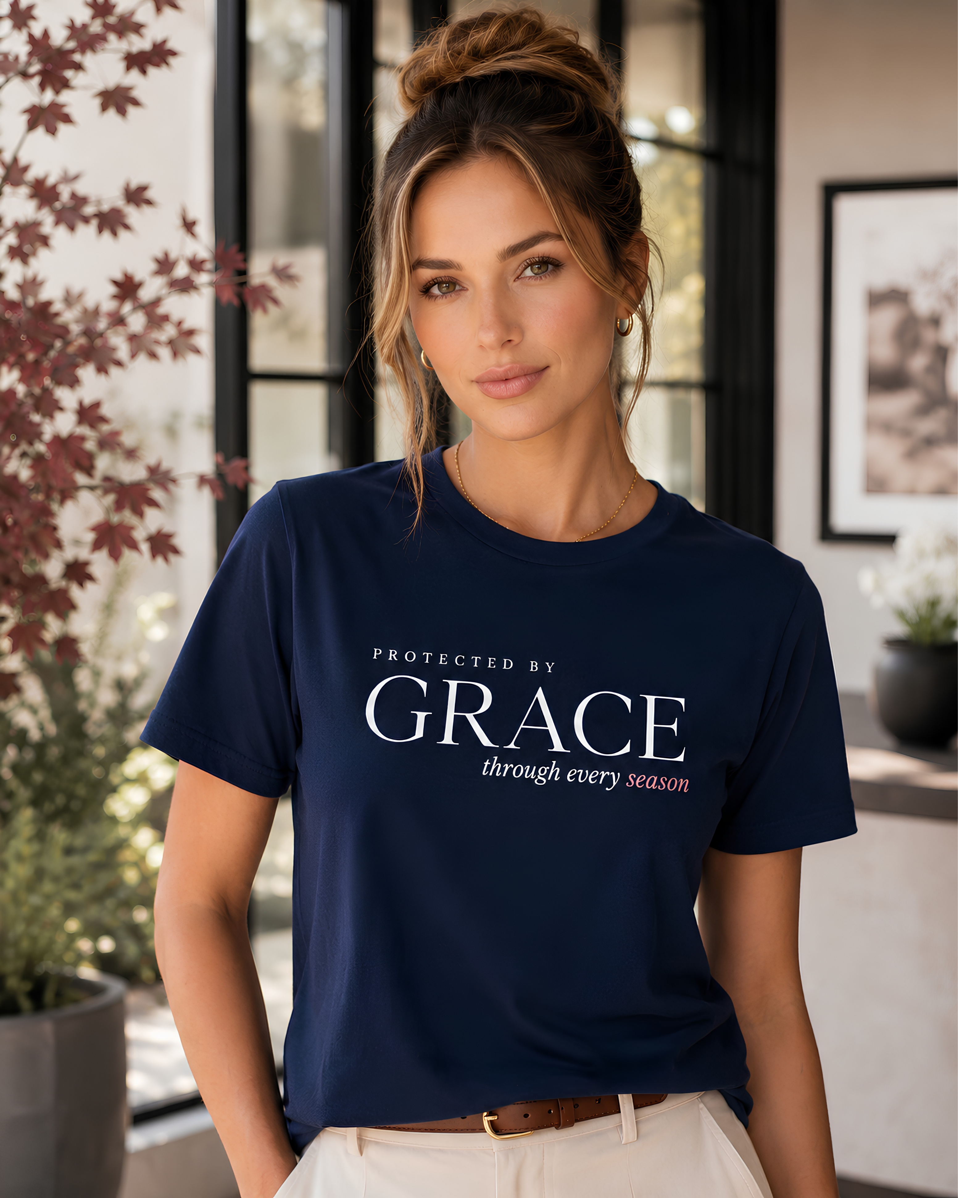 A woman wearing a navy blue Christian graphic t-shirt with the text 'PROTECTED BY GRACE through every season' in white and red, paired with cream pants, standing in a modern indoor setting with plants and windows.