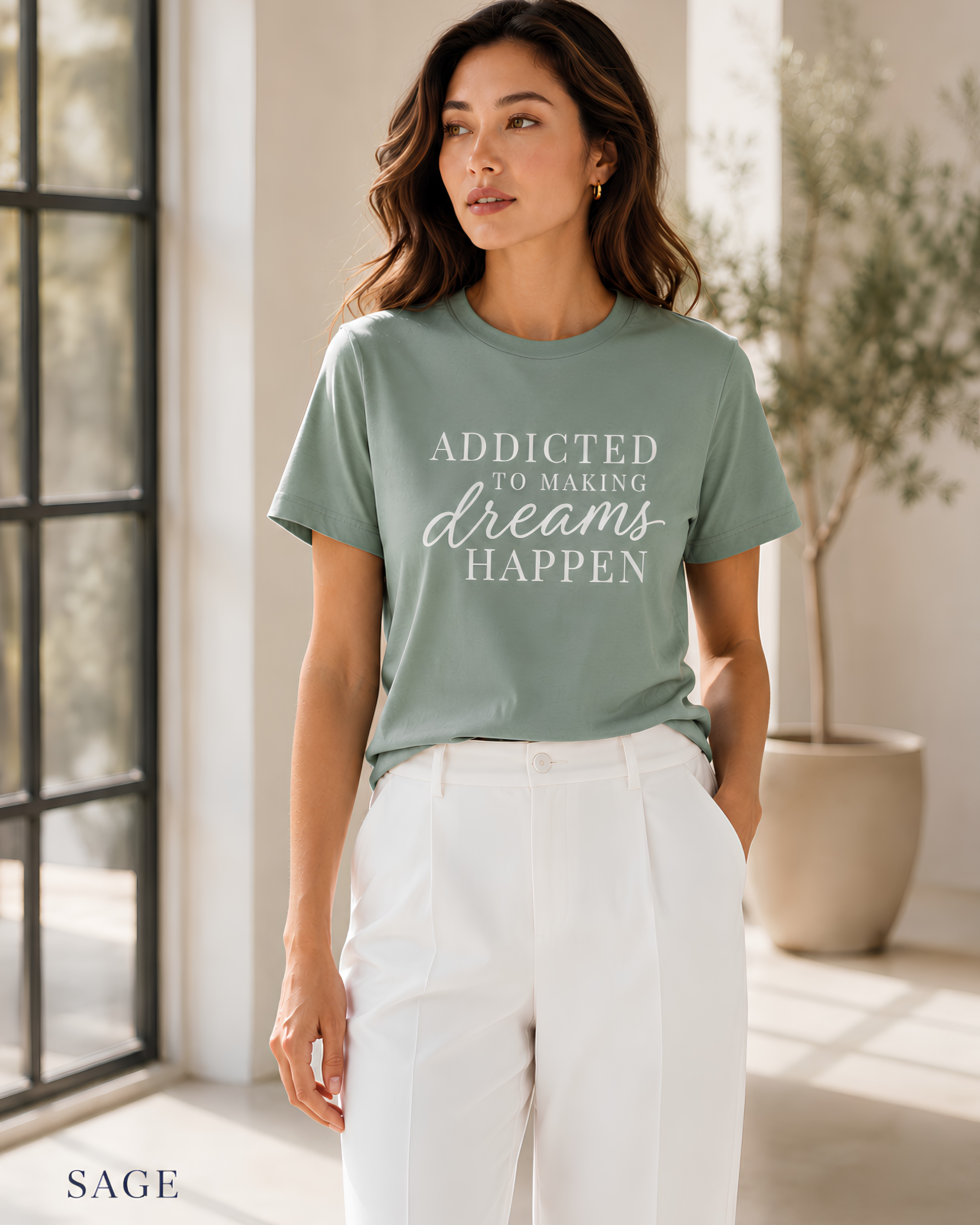 A woman wearing a sage green graphic t-shirt with the text 'ADDICTED TO MAKING dreams HAPPEN' in white, paired with white pants, standing in a bright, modern room with a window and potted plant.