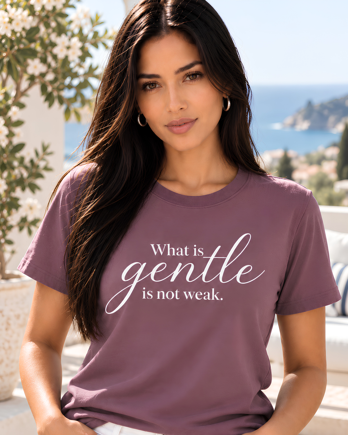 Young woman wearing a mauve What Is Gentle Is Not Weak inspirational graphic tee with white jeans on a bright Mediterranean-style terrace.