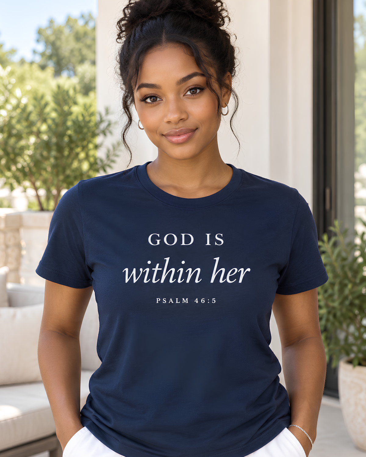 Young woman wearing a navy God Is Within Her Psalm 46:5 Christian graphic tee on a bright outdoor home terrace.