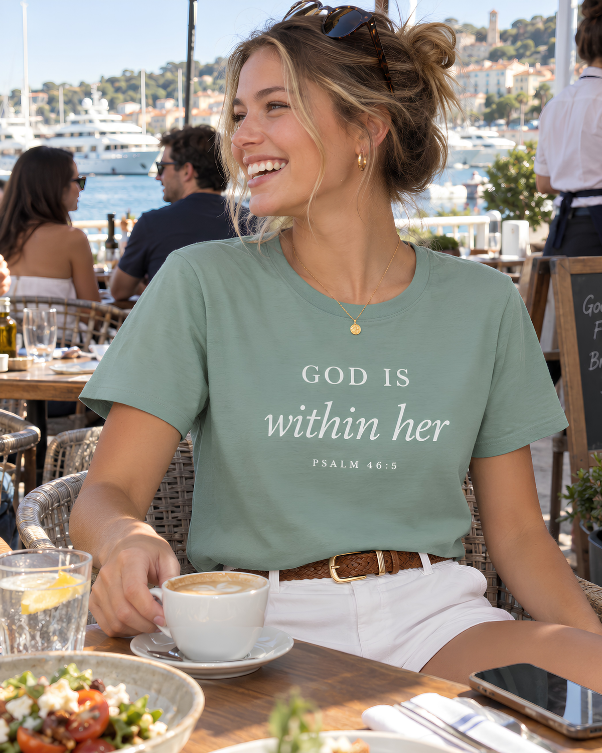 Young woman wearing a sage God Is Within Her Psalm 46:5 Christian graphic tee with white shorts at a sunny waterfront café.